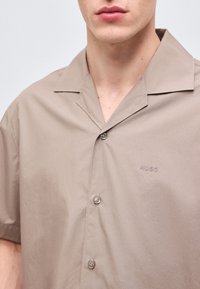 Gros plan d'une personne portant une chemise beige à manches courtes, boutonnée, avec un petit logo Hugo sur la poitrine gauche.