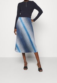 Kvinna iklädd en blå zigzag-mönstrad midiklänning, marinblå långärmad topp med dragkedjedetalj och svarta öppna klacksandaler.