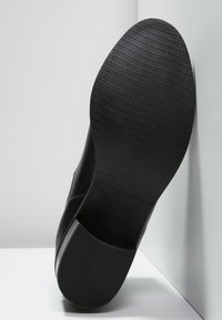 Czarna podeszwa buta z teksturowanej gumy, o zakrzywionym kształcie i tłoczonym znakiem rozmiaru. Projekt charakteryzuje się gładką górną krawędzią przechodzącą do podeszwy.