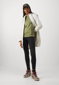 Lättviktig vit regnjacka, olivgrön t-shirt med ficka, svarta leggings, bruna och grå vandringskängor med röda accenter, grön keps med logotyp.