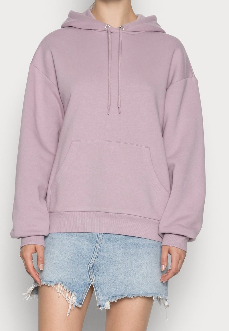 Personne portant un sweat à capuche oversize violet clair avec poche avant et cordons, associé à une mini-jupe en jean bleu clair effilochée.