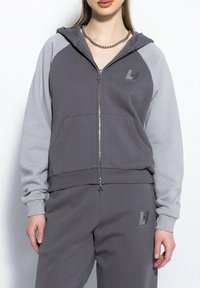 Kobieta ubrana w dwukolorowy szary hoodie na zamek i pasujące spodnie dresowe z logo na piersi i udzie, stojąca na białym tle.