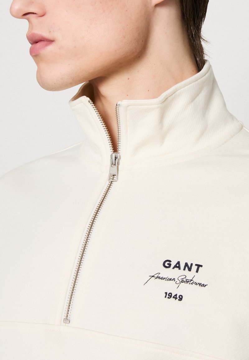 Pullover color crema con zip a metà, caratterizzato da un tessuto testurizzato e logo ricamato in blu navy. La cerniera è in metallo.