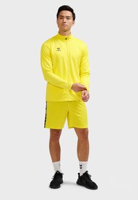 Gul atletikjakke med høj krave, lynlås og logo; matchende gule shorts med sorte striber i siden; kombineret med sorte sneakers og hvide sokker.