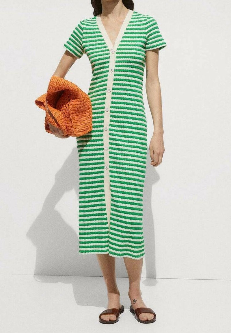 Femme portant une robe midi rayée verte et blanche à boutons, tenant un sac tissé orange, et portant des sandales plates marron.