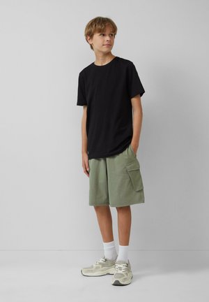 Schwarzes Baumwoll-T-Shirt, lockere Passform. Hellgrüne Cargo-Shorts mit Seitentasche. Graue und cremefarbene Sneaker, weiße Knöchelsocken. Neutraler Hintergrund.