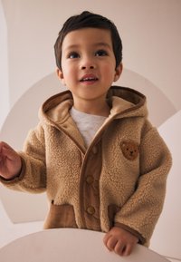 Veste en polaire beige avec une capuche brune, ornée d'un patch en forme d'ours, de boutons à l'avant et d'une surface texturée. Un enfant sourit, debout près d'un décor circulaire.