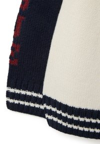 Suéter de punto con un diseño de bloques de color crema y navy, con letras rojas y puños de canalé a rayas blancas. Textura suave, grosor medio.