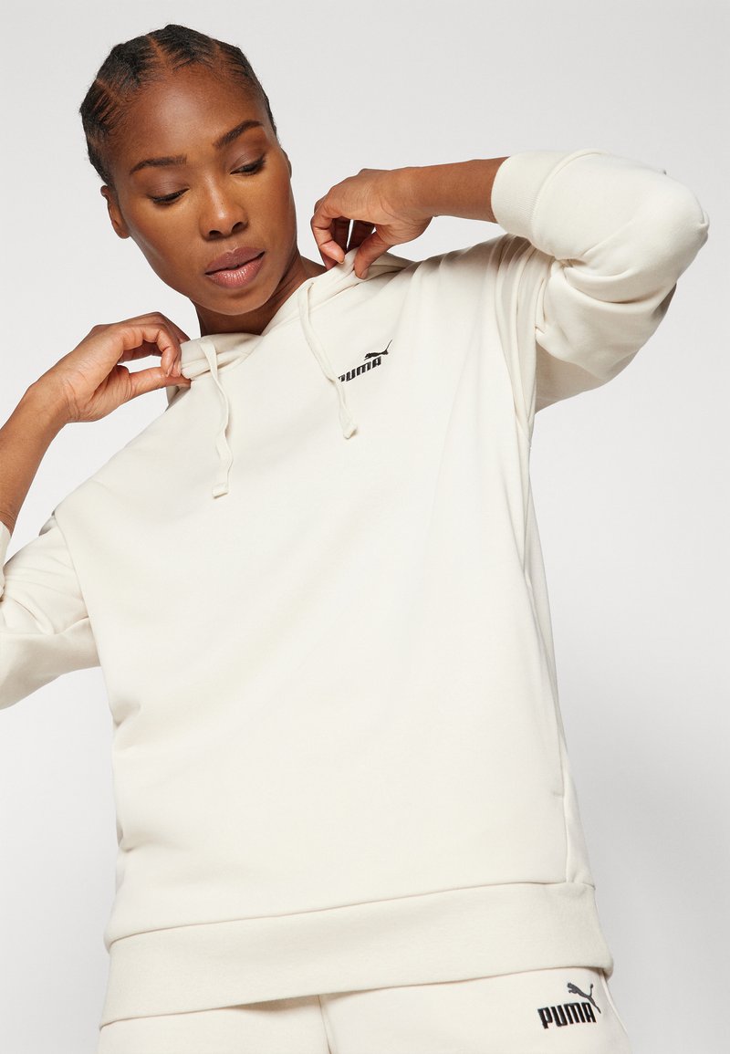 Cremfarbener Hoodie mit Kängurutasche, verstellbarem Kordelzug an der Kapuze, gerippten Bündchen und Saum. Vorne befindet sich ein kleines schwarzes Puma-Logo auf der Brust.