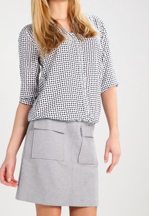 Femme blonde portant un chemisier blanc avec motif noir et une jupe gris clair dotée de deux poches à rabat à l'avant.