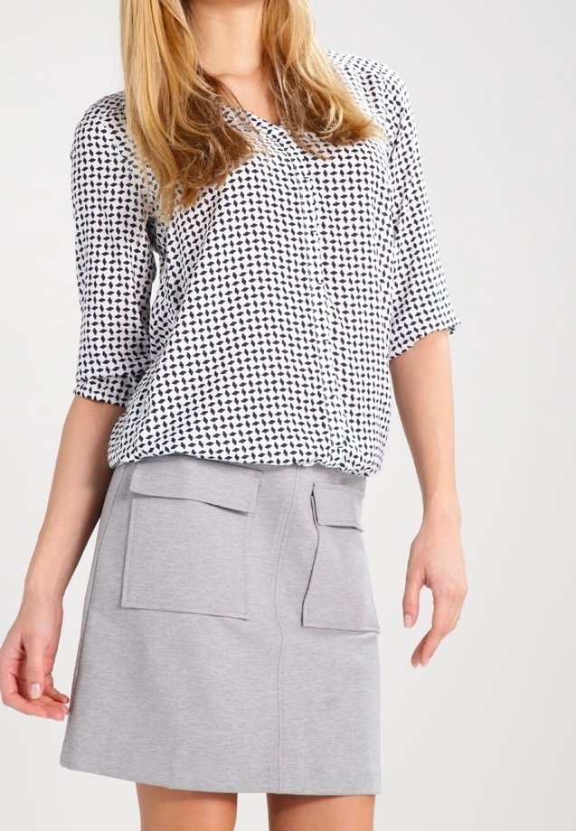 Femme blonde portant un chemisier blanc avec motif noir et une jupe gris clair dotée de deux poches à rabat à l'avant.