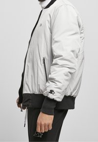 Zijaanzicht van een persoon die een witte bomberjack met ritssluiting draagt, met zwarte manchetten en een zwarte broek, beide voorzien van sterlogo's op de mouwen en het been.