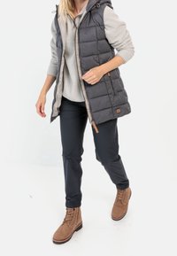 Dunkelgraue Steppweste mit Kapuze. Kombiniert mit einem hellgrauen Fleece-Oberteil, dunklen Hosen und braunen Schnürstiefeln.