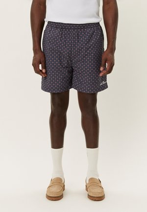 Mand iført marineblå mønstrede shorts, hvide sokker, beige loafers og en hvid ærmeløs top, stående mod en ensfarvet baggrund.