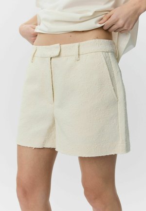 Zbliżenie na osobę noszącą kremowe, fasonowane szorty z teksturą, z przednimi kieszeniami i paskiem w talii, poprawiającą swój biały top.
