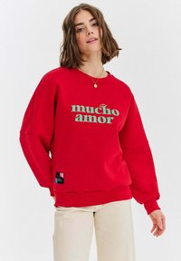 Czerwony sweter z luźnym krojem, szerokimi rękawami i napisem "mucho amor" w kolorze zielonym. Posiada czarną metkę na dolnym brzegu. Gładka tekstura.