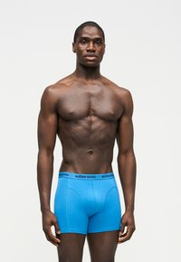 Μυώδης άνδρας στέκεται, φορά φωτεινά μπλε εσώρουχα μποξέρ Björn Borg, μπροστά σε απλό ανοιχτό φόντο.