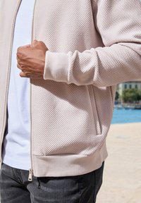 Hellrosa Zip-Up-Jacke aus strukturiertem Stoff; mit gerippten Bündchen und Saum. Getragen über einem weißen Hemd, kombiniert mit dunklen Jeans.