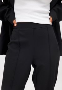 Črne elegantne hlače z gladko, mat teksturo, ki imajo oprijet pas in subtilne stranske šive. Nosi se jih z belim rebrastim zgornjim delom.