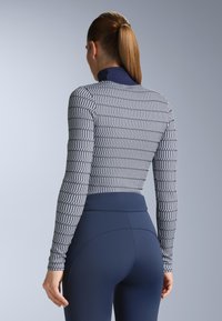 Sportowa bluzka z długim rękawem o geometrycznym wzorze w odcieniach szarości i granatu, z wysokim kołnierzem i dopasowanym krojem, zestawiona z granatowymi legginsami z wysokim stanem.