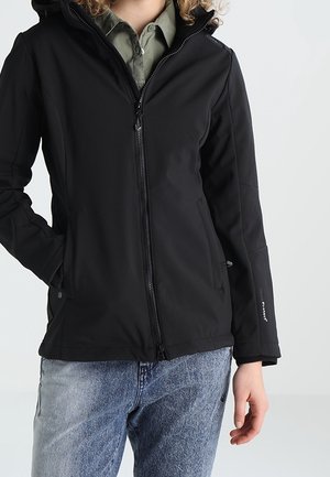 Personne portant une veste noire zippée sur une chemise verte à col et un jean bleu, debout avec une main dans la poche.