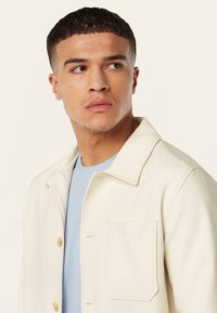 Lichtbeige jack met een klassieke kraag, twee knopen en een voorklep, gedragen over een blauwe shirt. Glad stofoppervlak.