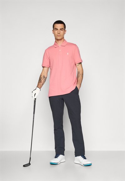 Polo shirt rosa claro, calças pretas, sapatos brancos com detalhes azuis, segurando um taco de golfe, tatuagens visíveis no braço esquerdo.