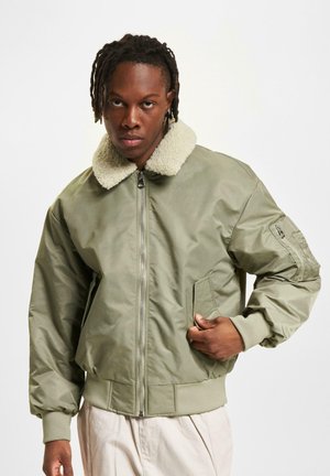 Homme avec des dreadlocks portant une veste bomber vert clair avec un col en fourrure blanche, un pantalon beige, se tenant devant un fond blanc uni.