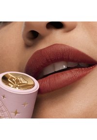 Matte rødbrune læber viser detaljeret tekstur; et pink læbestift rør med et gyldent segl og præget design holdes tæt på.