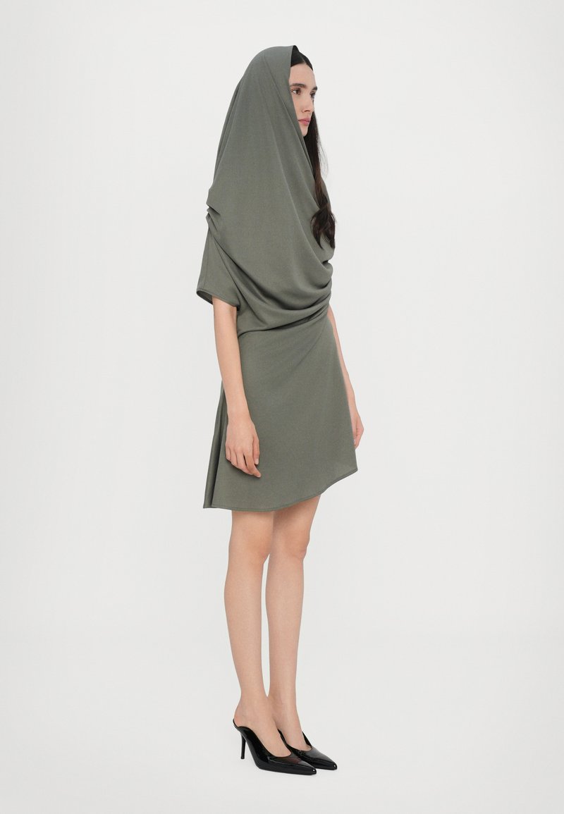 Asymmetrical olive green dress with draped hood, short sleeves, and soft texture, paired with black high-heeled shoes.