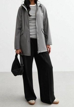 Femme portant une veste à capuche grise par-dessus une chemise rayée noire et blanche, un pantalon noir à jambes larges, des chaussures beiges, et tenant un petit sac à main noir.