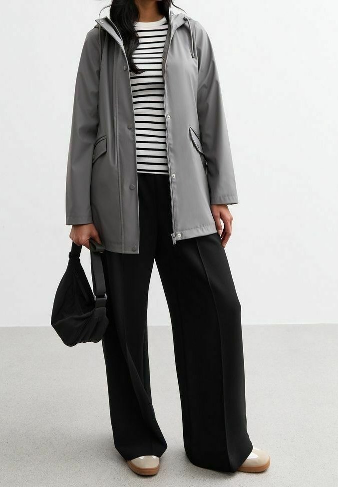 Femme portant une veste à capuche grise par-dessus une chemise rayée noire et blanche, un pantalon noir à jambes larges, des chaussures beiges, et tenant un petit sac à main noir.