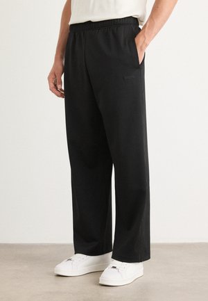 Black, relaxed-fit trousers with an elastic waistband and pockets, made of a smooth, lightweight fabric, paired with white sneakers.