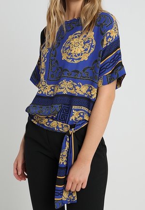 Top blu con motivo barocco dorato, maniche corte e dettaglio in vita annodato. Il materiale appare liscio con una vestibilità ampia e rilassata.