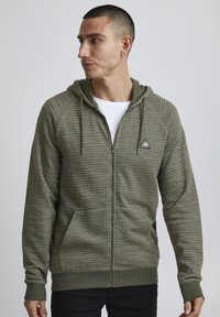 Haut à capuche zippé olive avec un motif texturé, une capuche, des poches avant et un petit logo. Poignets et ourlet côtelés. Matériau léger.