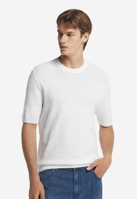 Kortärmad vit ribbad tröja med rund halsringning och en avslappnad passform, kombinerad med blå jeans. Inga synliga mönster eller metalldetaljer.