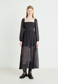 Czarna długa sukienka z długim rękawem i kwadratowym dekoltem, z koronką w kwiatowy wzór, przezroczystym materiałem i dopasowanym gorsetem, w zestawieniu z brązowymi botkami za kostkę.