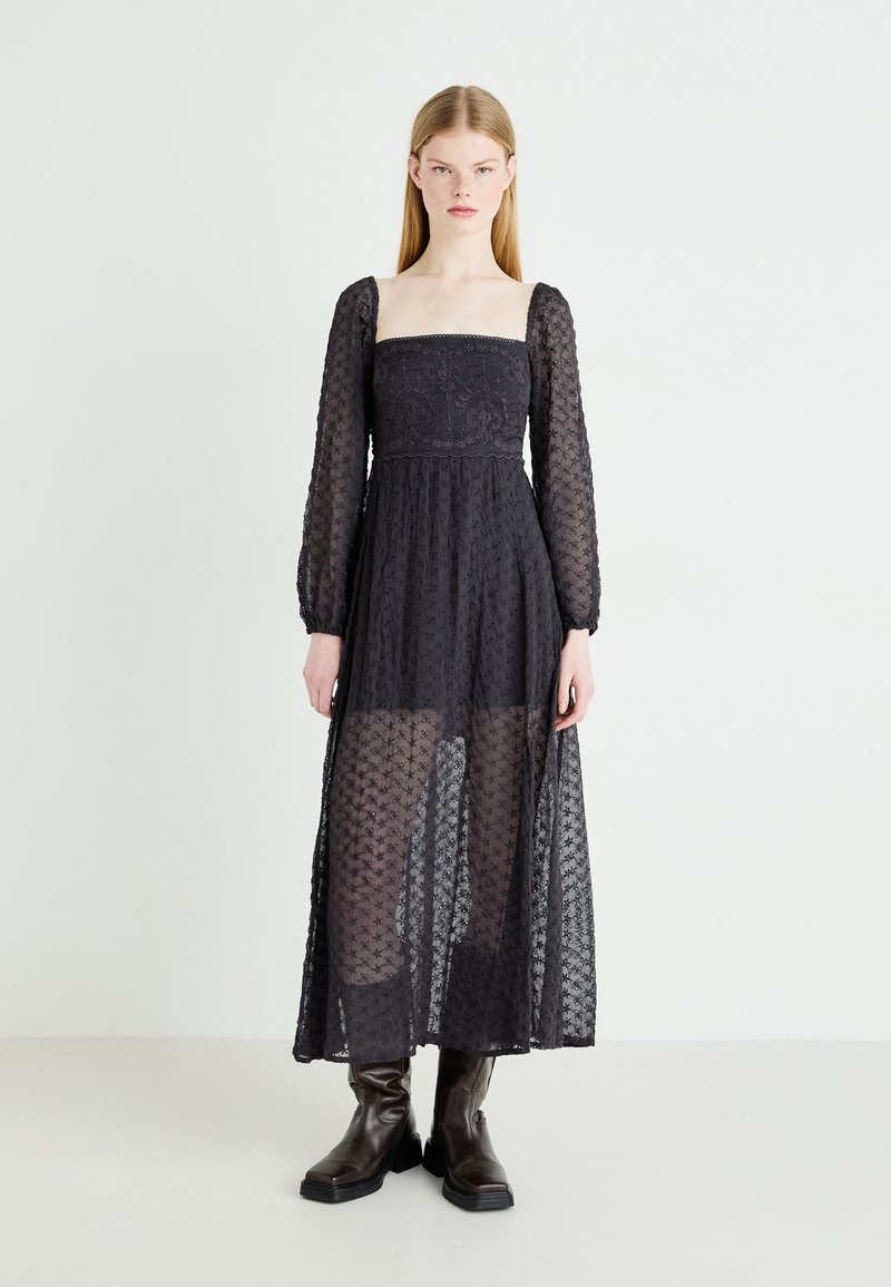 Czarna długa sukienka z długim rękawem i kwadratowym dekoltem, z koronką w kwiatowy wzór, przezroczystym materiałem i dopasowanym gorsetem, w zestawieniu z brązowymi botkami za kostkę.