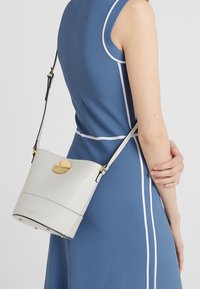 Borsa a tracolla in pelle bianca con texture, forma arrotondata, dettagli in oro e tracolla regolabile, indossata con un vestito blu.