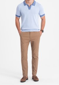 Mężczyzna ubrany w jasnoniebieską koszulkę polo z krótkim rękawem, beżowe dopasowane spodnie, brązowe zamszowe mokasyny z frędzlami i czarny zegarek na rękę, stojący z rękami w kieszeniach.