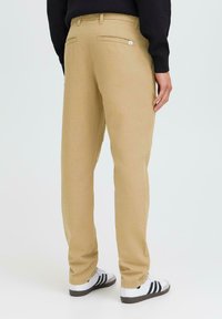 Pantalons en coton beige avec deux poches arrière, design à jambe droite et finition texturée, associés à des chaussures de sport noires et blanches.