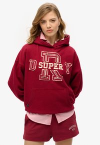 Bordeaux hoodie met grote crèmekleurige letters en een voorzak, gelaagd over een lichtroze shirt. Korte bordeauxbroekjes met merkdetail.