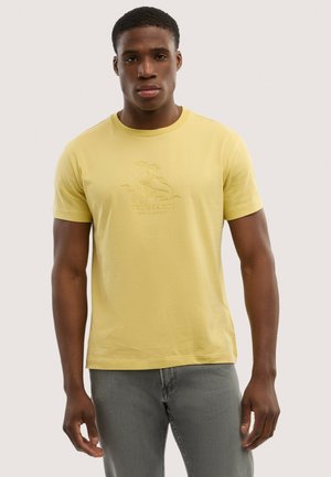 Uomo che indossa una T-shirt gialla Trussardi con logo in rilievo e jeans grigi, in piedi davanti a uno sfondo chiaro e semplice.