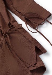 Manches en tissu marron texturé avec fermetures à nouer réglables et fentes aux poignets sur fond blanc.