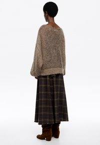 Osoba s krátkymi kučeravými vlasmi má na sebe prehnane veľký hnedý sveter, dlhú tmavo kockovanú sukňu a hnedé semišové čižmy, stojí so zady kom. 