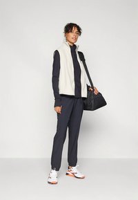 Lichtgewicht crèmekleurig fleecevest over een donkerblauw shirt met lange mouwen, gecombineerd met donkere joggers en witte sneakers met oranje accenten. Zwarte tas over de schouder gehangen.