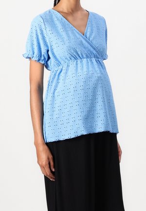 Femme portant un haut cache-cœur bleu clair à manches courtes ajouré, associé à une jupe noire, vue du buste jusqu'à mi-cuisse.