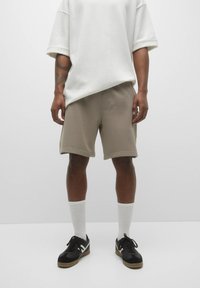 Shorts beige, matière douce, longueur genou, taille élastique, assortis à un t-shirt blanc ample et des baskets noires avec des chaussettes blanches.
