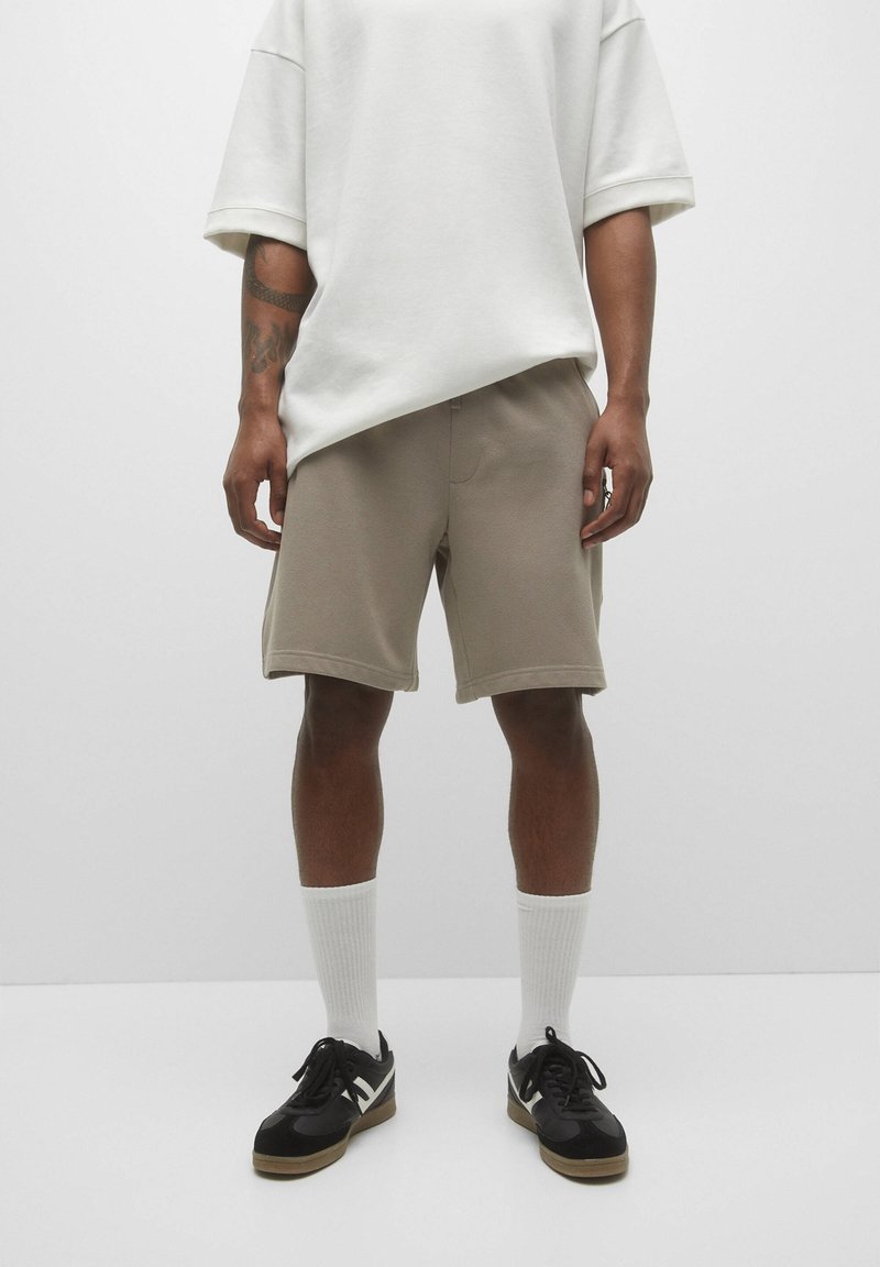 Shorts beige, matière douce, longueur genou, taille élastique, assortis à un t-shirt blanc ample et des baskets noires avec des chaussettes blanches.