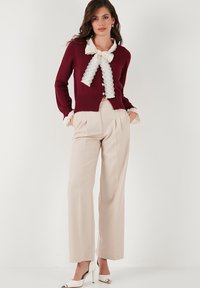 Burgundský svetr s krajkovým límcem a detailem na mašli, kombinovaný s přizpůsobenými béžovými kalhotami. Má přízí zakončení a knoflíkové zapínání.
